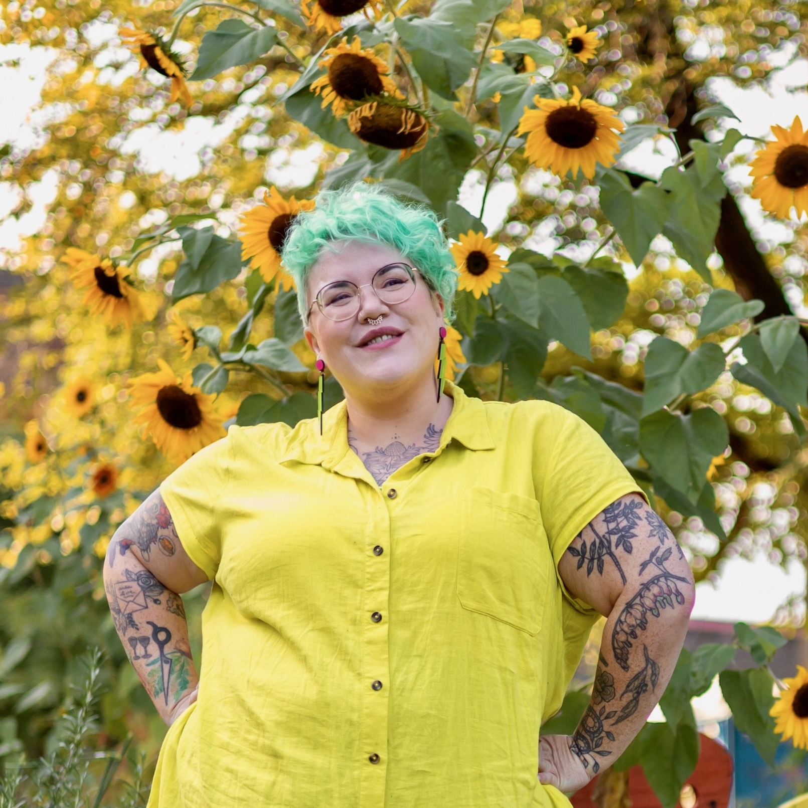 Hannah, with her hands on her hips, smiling in front of a sunflower tree. 