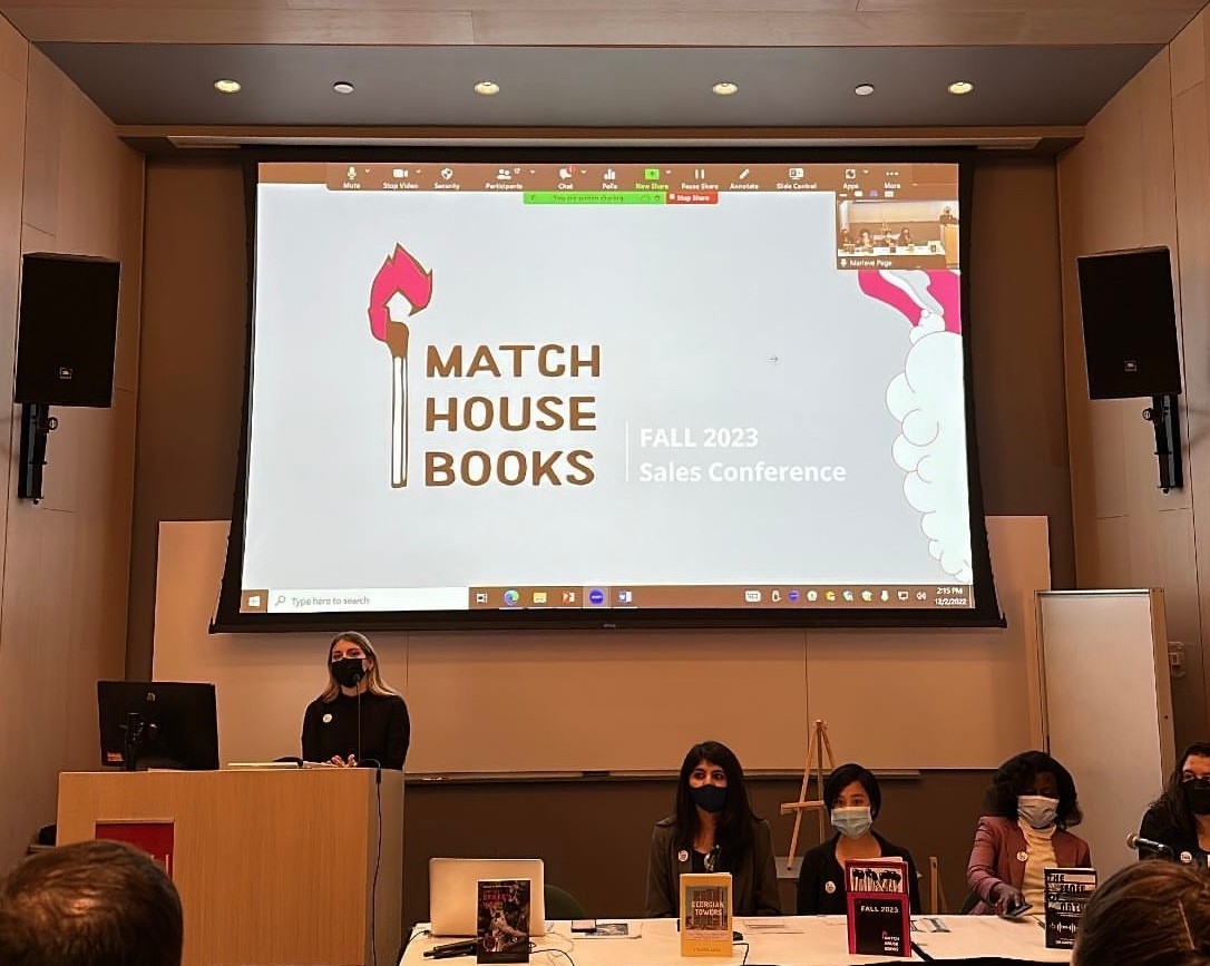 Five SFU Master of Publishing (MPub) students presenting (behind them) the cover of their group’s press catalogue:  A (pink) lit white and brown matchstick. Next to it is the Press’s name, MATCH HOUSE BOOKS in bold. On the right side of the company logo,  a text reads “Fall 2023, Sales Conference”. 
