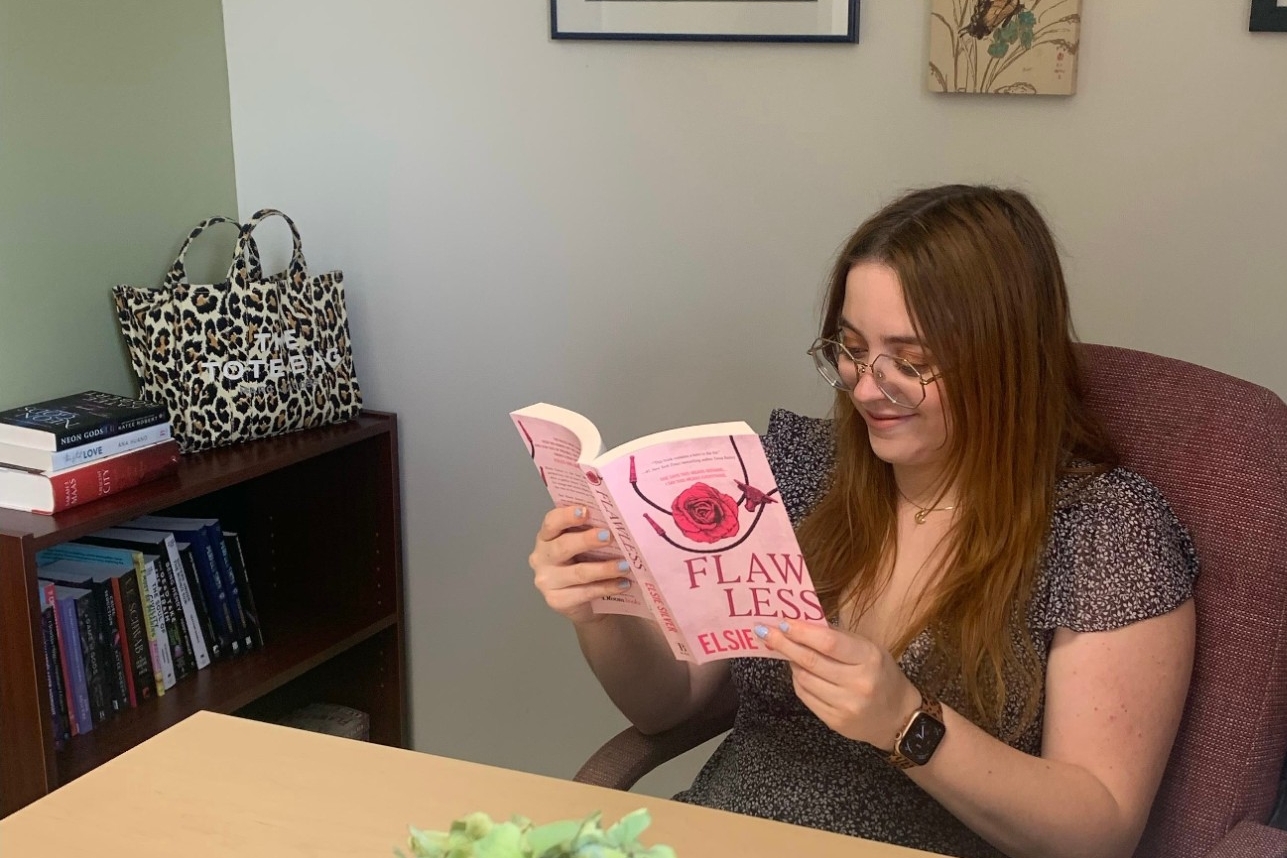 Current Master of Publishing student Christina Morden smiles as she sits in her office, holding a book titled Flawless, a small-town romance novel and TikTok bestseller by Elsie Silver. 