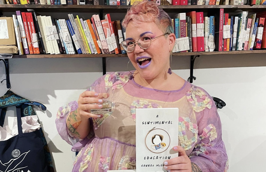Hannah McGregor holding a drink on her right hand and her newly published book, A Sentimental Education, on the other, as she poses in front of a book shelf at a local indigenous book store called Iron Dog Books.
