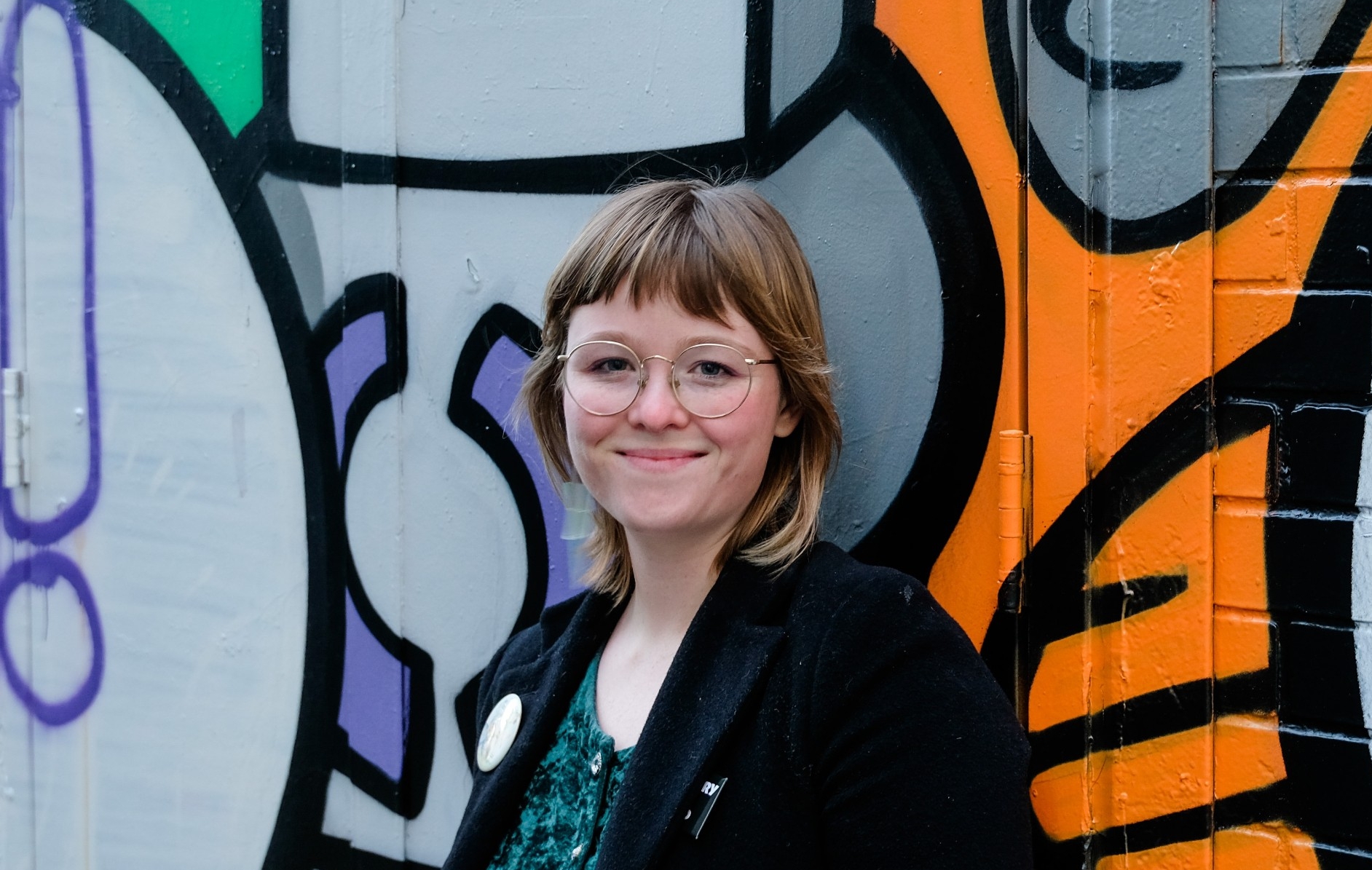 Master of Publishing student Zoe Mix smiles as she poses in front of a colourful and vibrant graffiti wall. She is wearing glasses and is donning a black jacket over a dark green velvet top. 