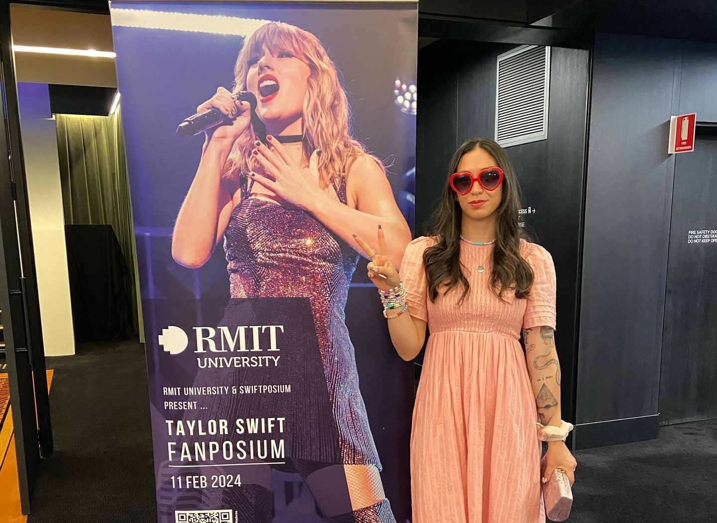 Jarin, in a peach dress and bright pink heart-shaped sunglasses, poses beside a standee of Taylor Swift at the Swiftposium event held in Melbourne, Australia. An overlay text reads (in bold): RMIT University. RMIT University and Switposium present Taylor Swift Fanposium, 11 Feb, 2023. Under the text is a QR code, as well as logos of the sponsors of the event. 