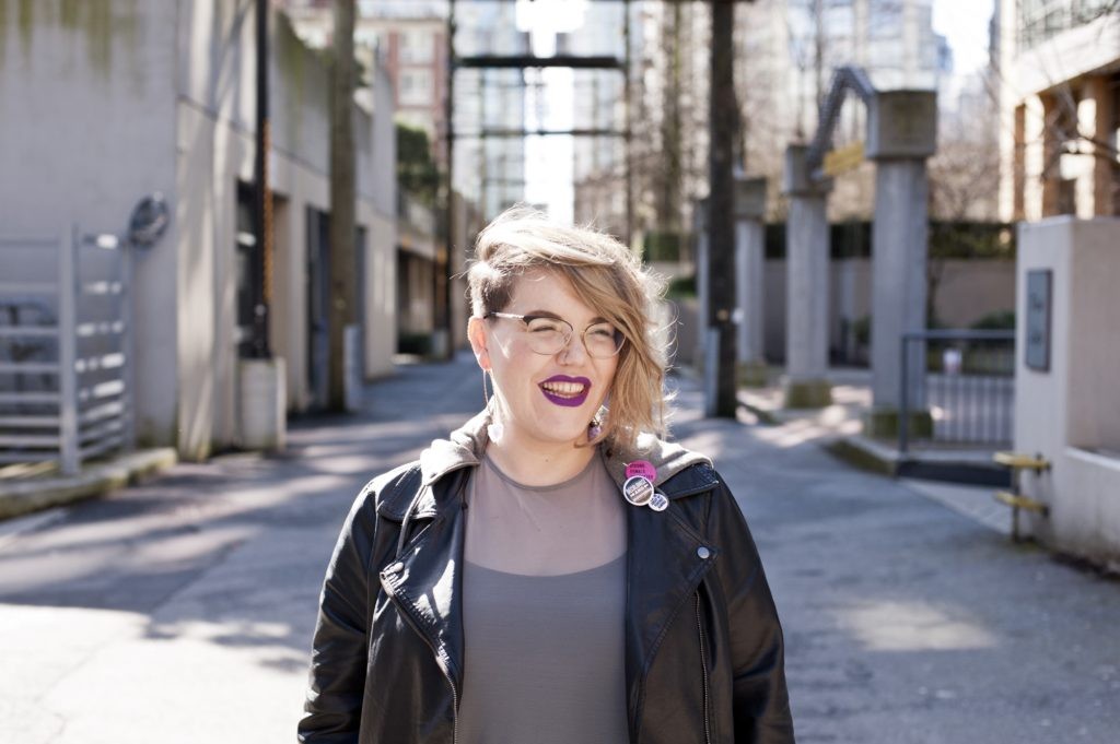 A smiling woman stands in an alley, wearing a leather jacket, short hair and purple lipstick