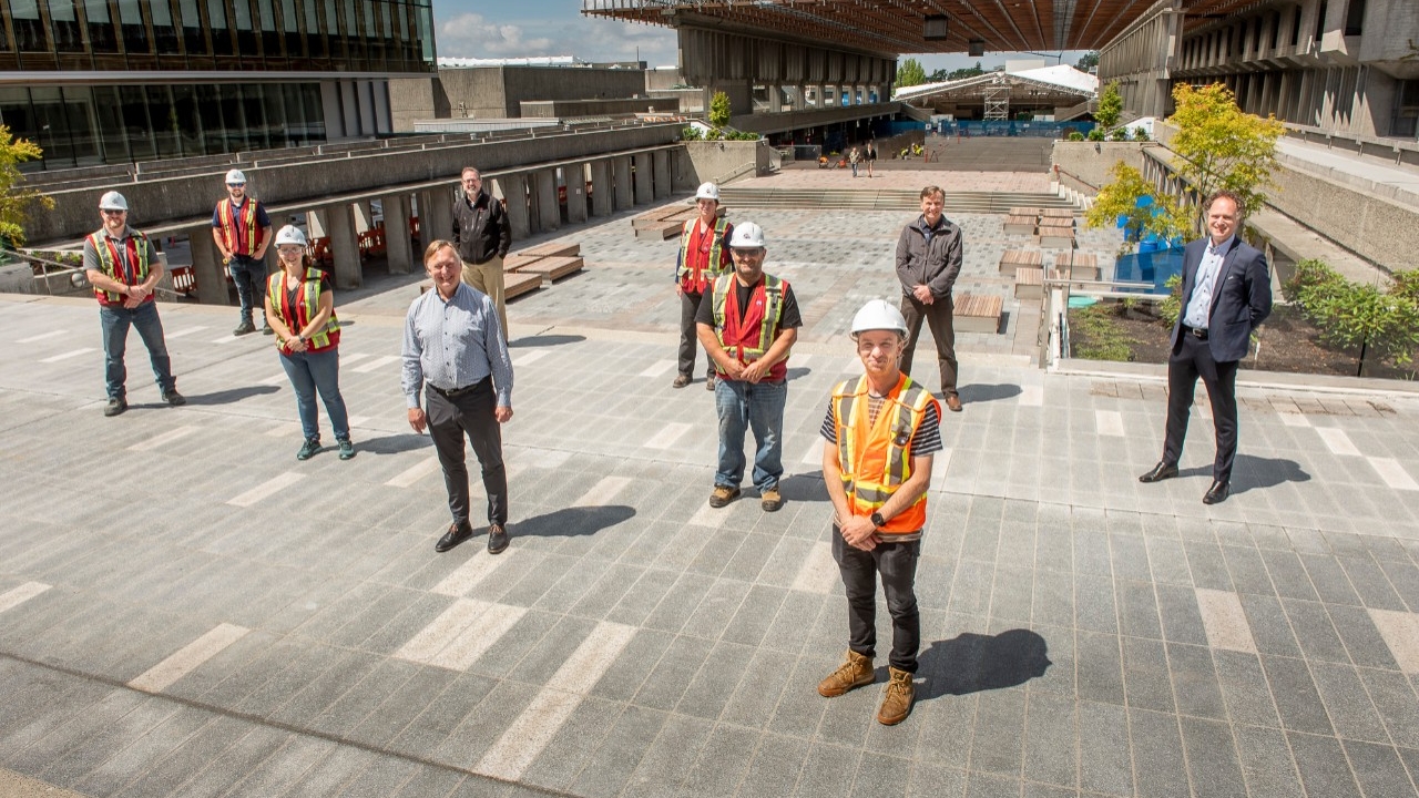 10 members of the plaza renewal team, many in construction gear, pose in the AQ on a sunny day