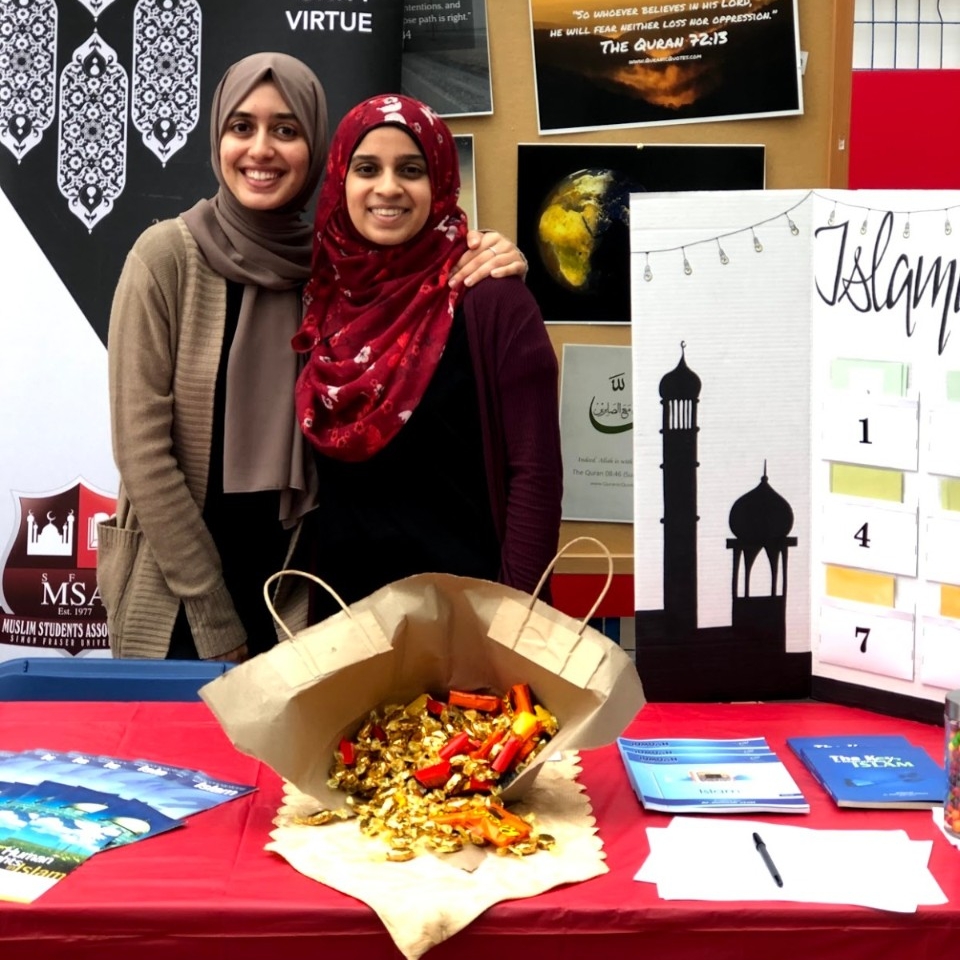 Two women stand behind a table with a bag of candy and a posterboard on it. Tala Adlouni wears a brown hijab and cardigan, and Rasha Syed wears a red hijab with flowers on it and a red caridgan. The word "Islam" is visible on the posterboard but the rest is cut off by the photo.  