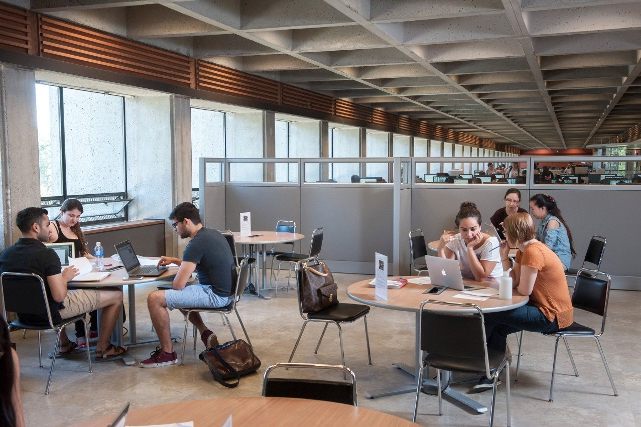 Inside SFU's W.A.C. Bennett Library, several students and peer educators work together in small groups at multiple tables, looking at laptops and papers.