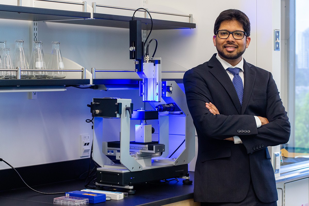 Sami Khan is standing in a lab and smiling at the camera with his arms crossed in front of his chest. He wears a black suit and blue striped tie. To his left is a futuristic-looking microscope. 