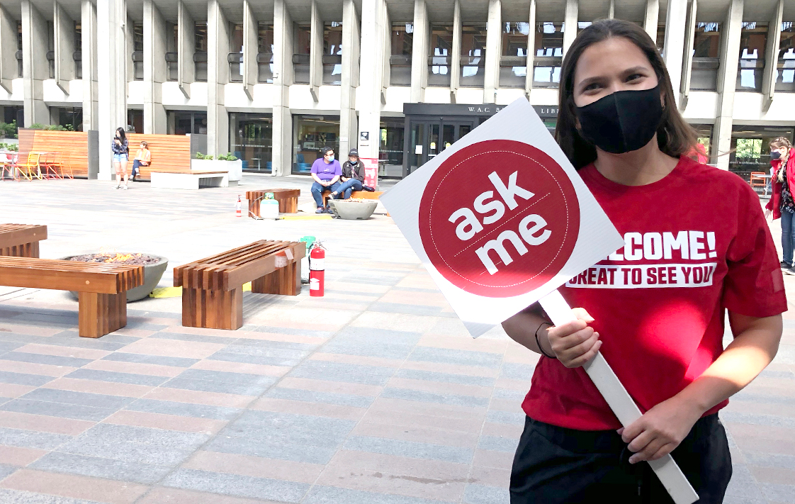 A "Ask Me" ambassador wearing a mask and holding up a sign saying, "Ask me"