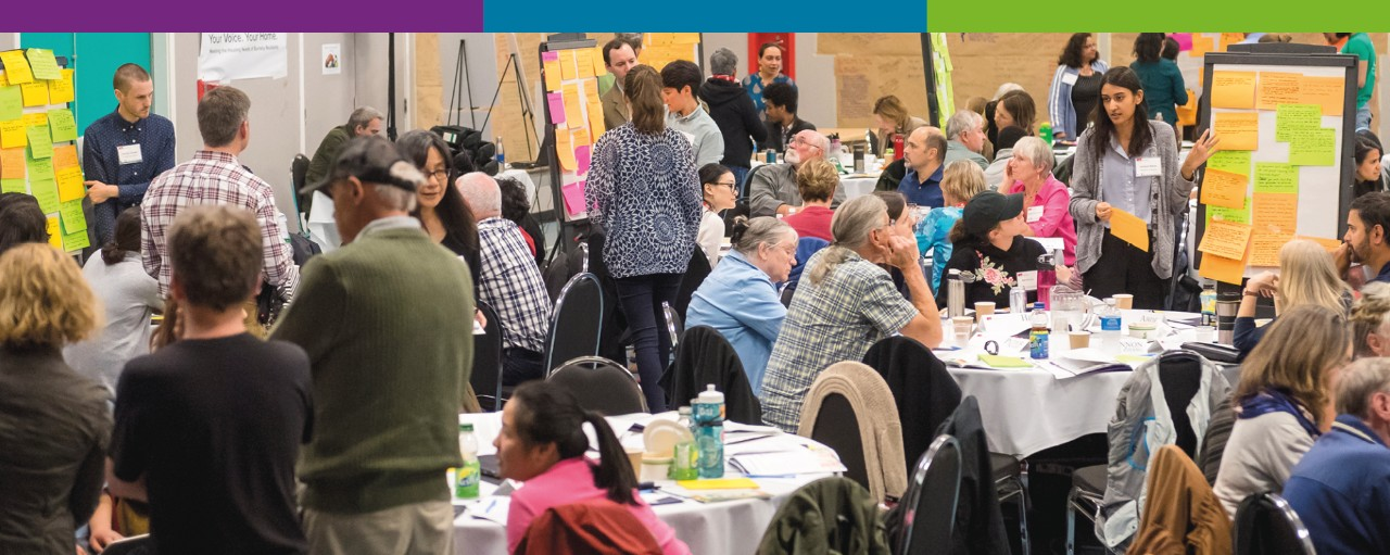 Panoramic photo of Burnaby residents in groups interacting with open house volunteers with sticky notes stuck to whiteboards.