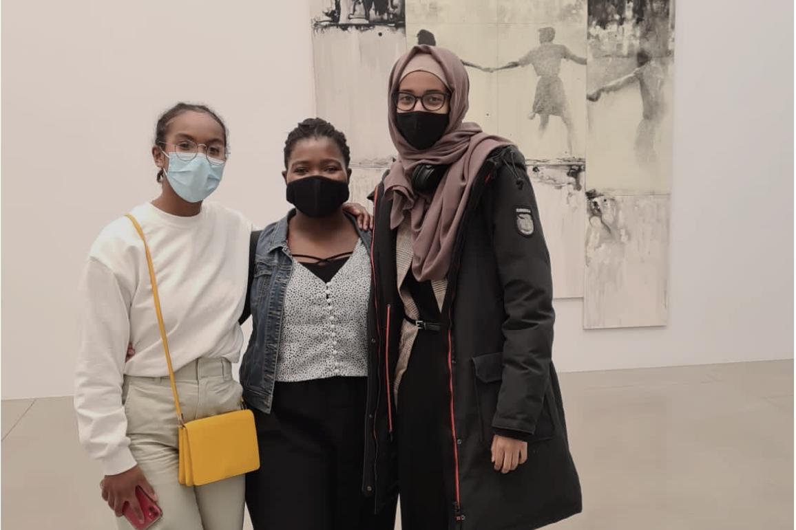 Three Black students stand in front of an abstract black-and-white piece of artwork, smiling behind their masks. Linda (left) wears a white shirt and bright yellow purse and has her arm around Sophonie (middle), who wears a grey sweater and jean jacket. Balqees (right) wears a brown hijab and long black jacket.