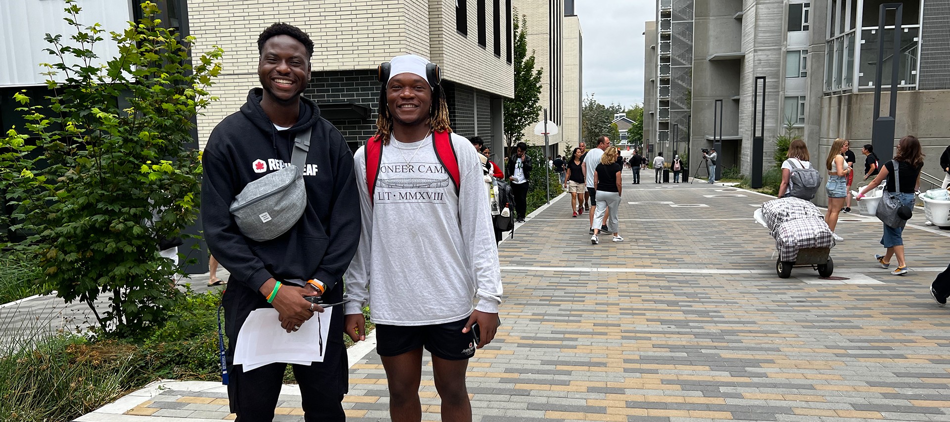 2 student athletes standing in the walkway between East Towers and North Towers during Move In, smiling in front of the camera on a sunny day