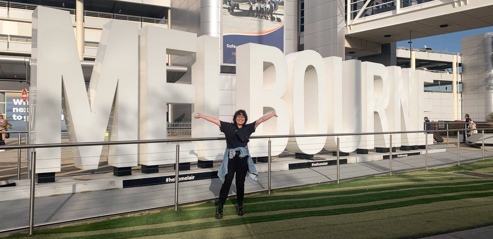 Junelle Knihniski upon arrival in Melbourne, Australia. Standing in front of large MELBOURNE sign at airport with outstretched arms overheard. 