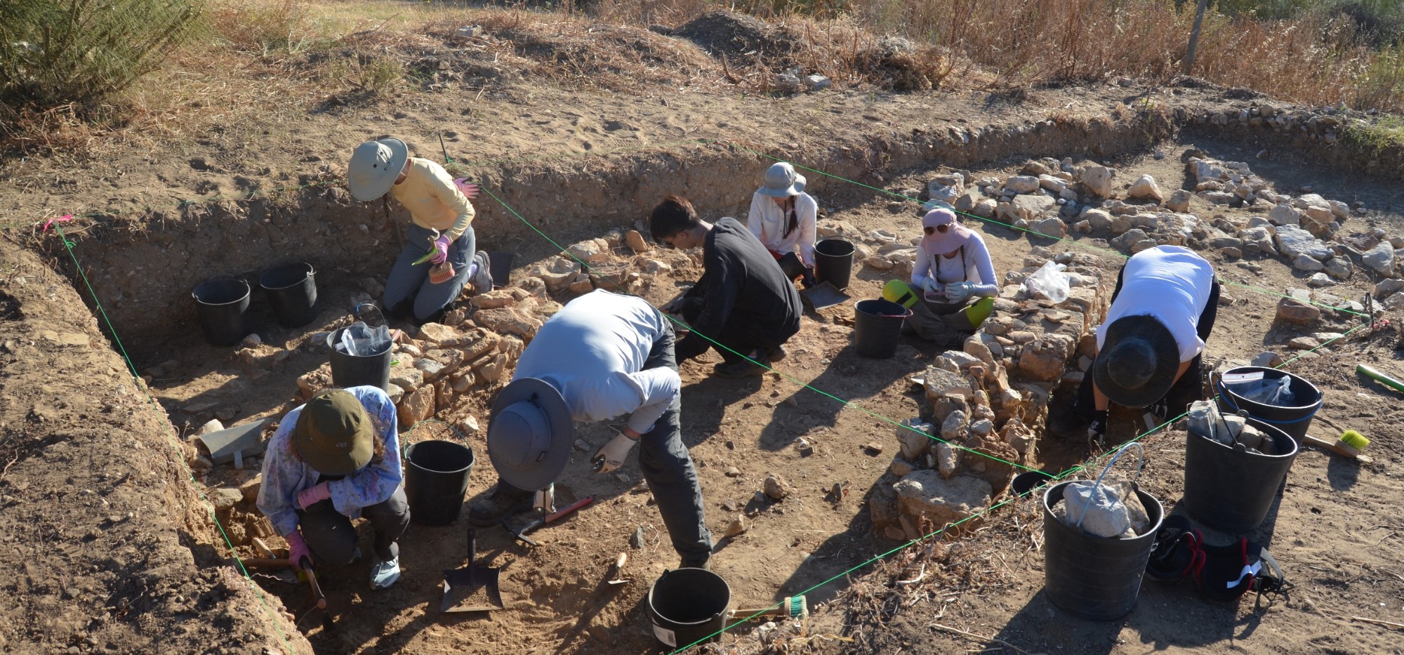 Seven SFU students, wearing wide brim hats, using dig tools to excavate site at Cacala Velha Village, Algarve, Portugal
