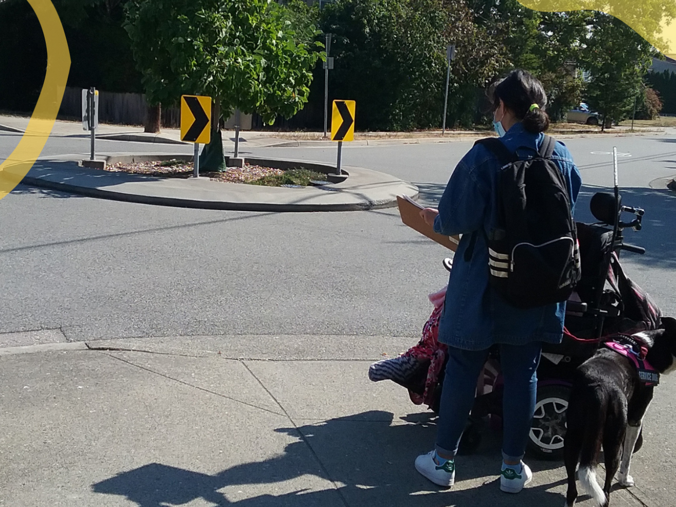 A person holding a clipboard standing next to a person in a wheelchair at a corner of the sidewalk. 