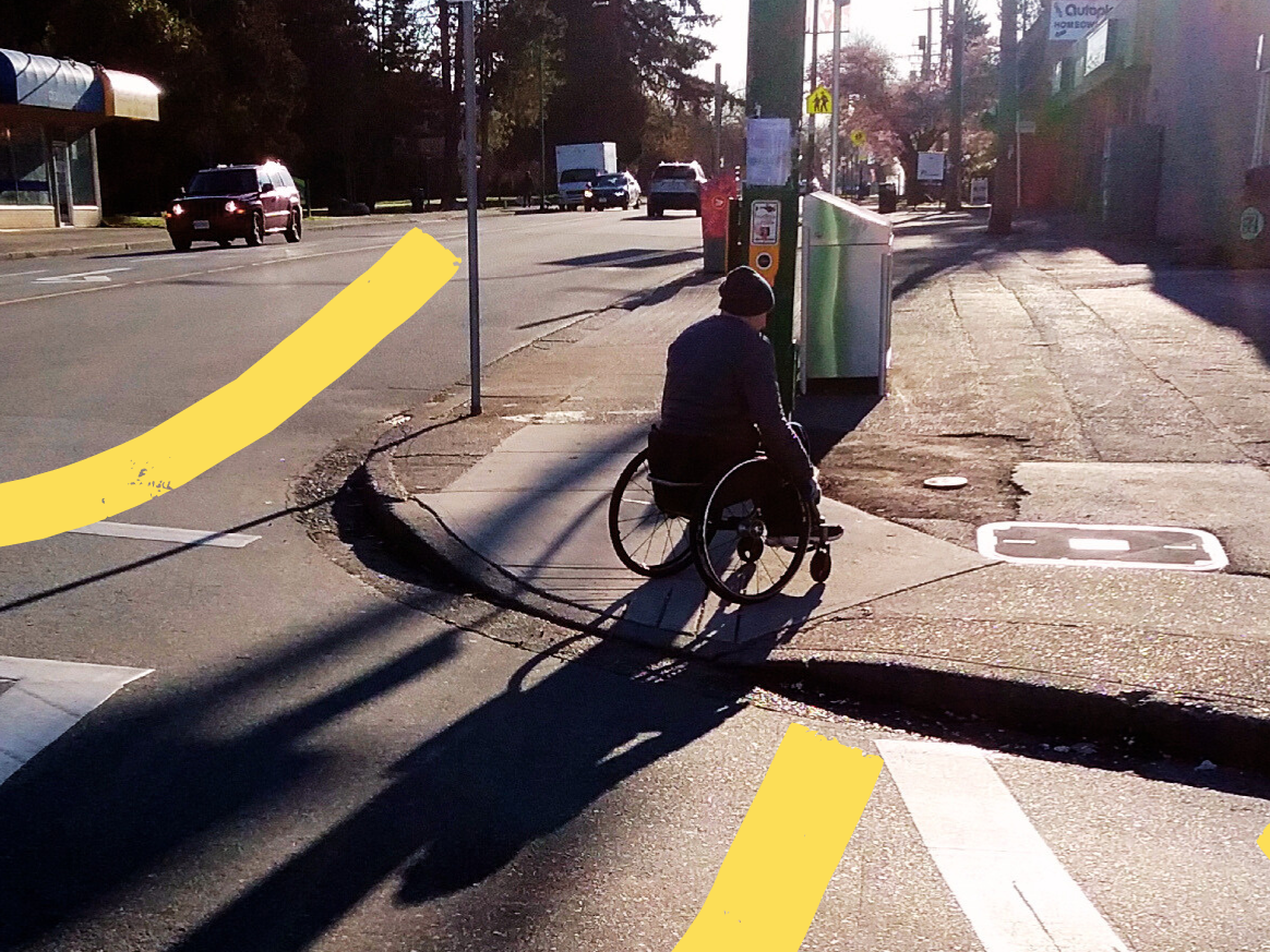 A person in a wheelchair wheeling up a curb ramp on the sidewalk. The curb ramp is aligned to to the middle of the intersection.  
