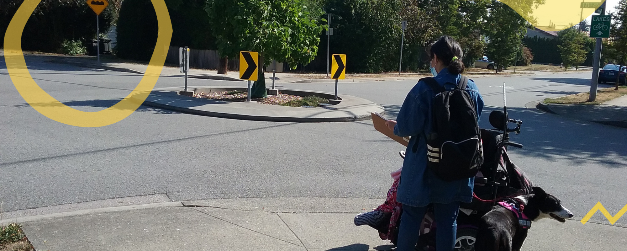 A research assistant holding a clipboard standing next to a person in a wheelchair at a corner of the sidewalk. 