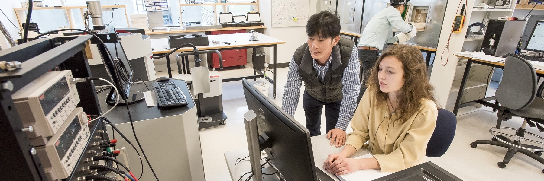 Two people in a lab working at a computer, with various types of machinery and technology around them in the room