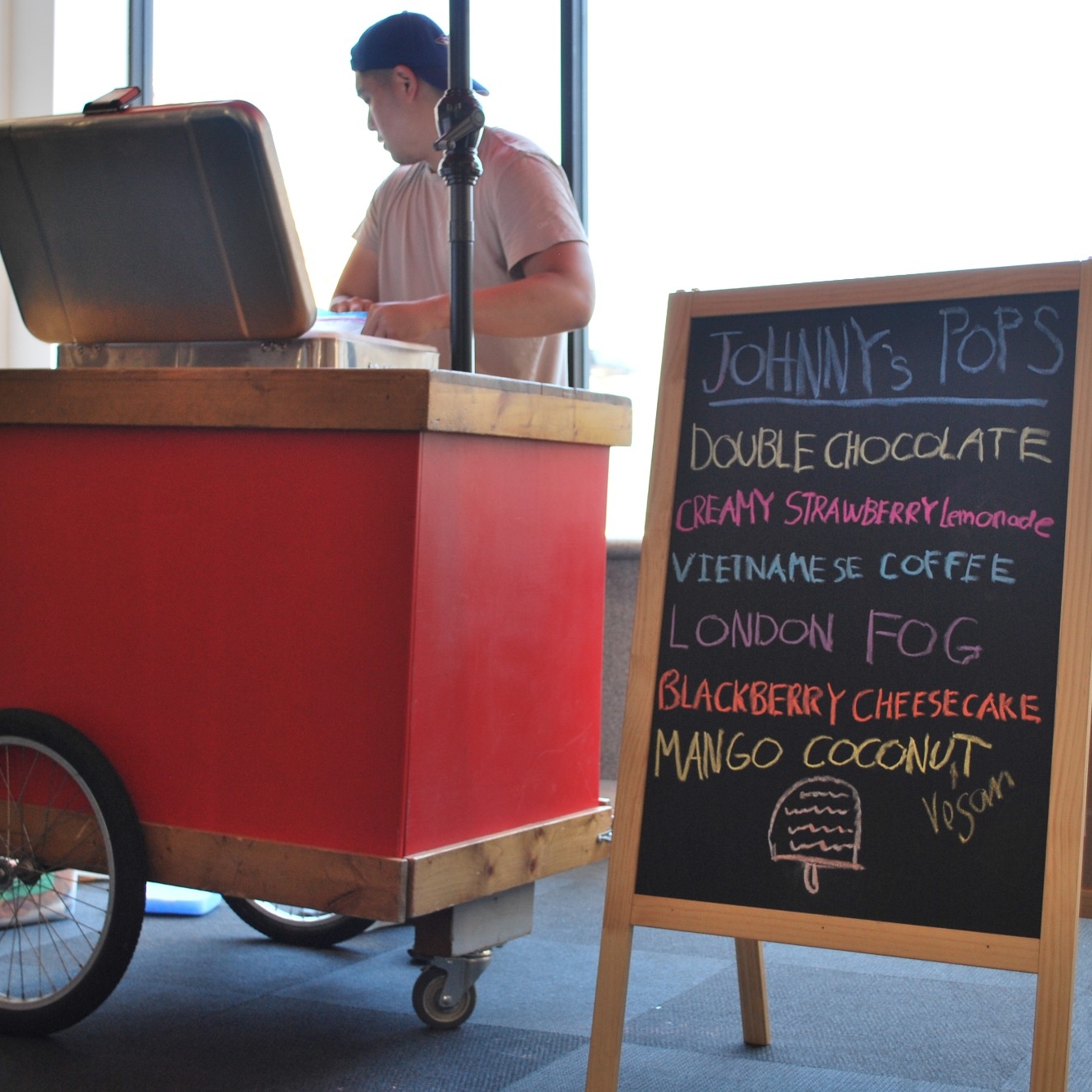 Johnny's Pops staff member standing behind a cart and sign that lists popsicle flavours available at the 2022 Summer Social